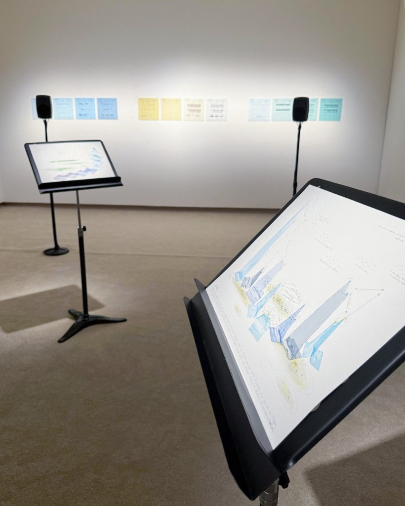 A hand-drawn score on a music stand in the foreground: layered blue line drawings of sound shapes, yellow watercolour contours and handwritten notes. Behind it, a second score on a stand, two speakers and a row of twelve blue, yellow and green analysis prints on a lit white wall.
